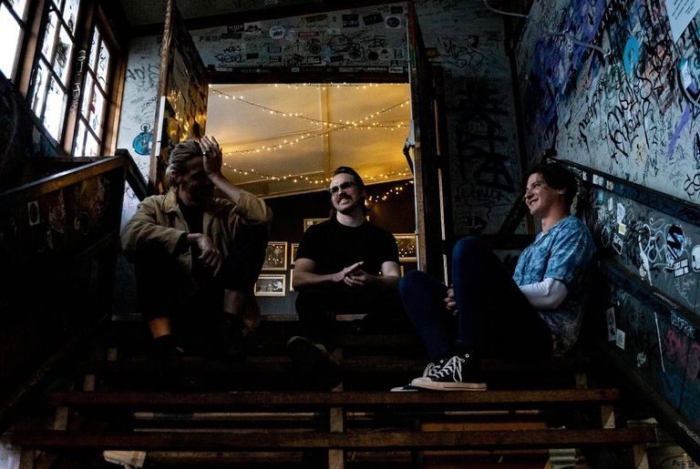 Brisbane indie rock band Bugs sitting on a graffiti-covered staircase inside a music venue, laughing together during a promo shot.